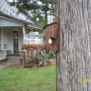 May include: A wooden bird feeder attached to a tree trunk. The feeder has a clear glass jar for holding birdseed and a wooden platform for birds to perch on. The feeder is mounted on a wooden bracket and is attached to the tree with screws. The tree trunk is brown and has a rough texture. The date 03/08/2018 is written on the tree trunk.