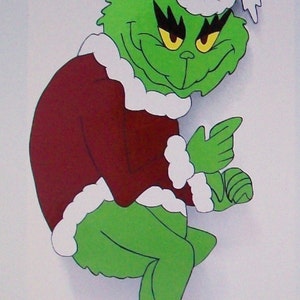 May include: A green cartoon character with a mischievous grin, wearing a red coat and hat trimmed with white fur. The character is posed with one finger pointing, and red boots. A festive holiday decoration.
