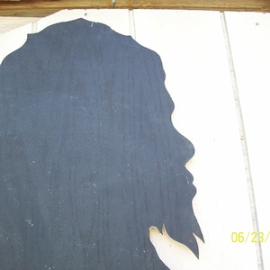 May include: A black silhouette of a person's head and shoulders, cut out of wood and mounted on a white wooden background. The silhouette is facing to the right, and the person's hair is long and flowing.