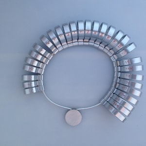 May include: A silver ring sizer with numbered rings from size 4 to size 15. The rings are attached to a metal band with a circular base.