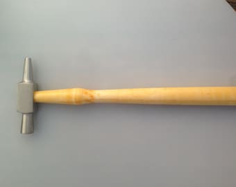 May include: A metal hammer with a light-coloured wooden handle. The hammer head is silver and has two different striking surfaces. The handle is smooth and appears to be made of a light-coloured wood. The hammer is designed for metalworking or similar tasks.