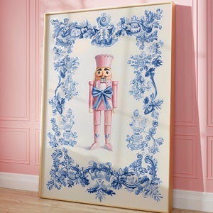 Puede incluir: Una impresión enmarcada con un soldado cascanueces rosa con un lazo azul, sobre un fondo blanco. El fondo está enmarcado por un diseño floral azul. El marco es de color dorado.