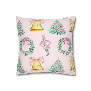 May include: A pink throw pillow with a repeating pattern of Christmas trees, wreaths, bells, and a nutcracker. The trees are green with red and gold ornaments. The wreaths are green with red bows. The bells are gold with red bows. The nutcracker is wearing a pink and green uniform.