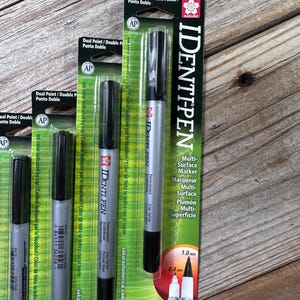 Puede incluir: Varios marcadores multi-superficie IDENTI-PEN negros y plateados se muestran en su embalaje original. El embalaje es verde y blanco, con el nombre de la marca y los detalles del producto. Los marcadores tienen puntas dobles, una de 1,0 mm y otra de 0,4 mm.