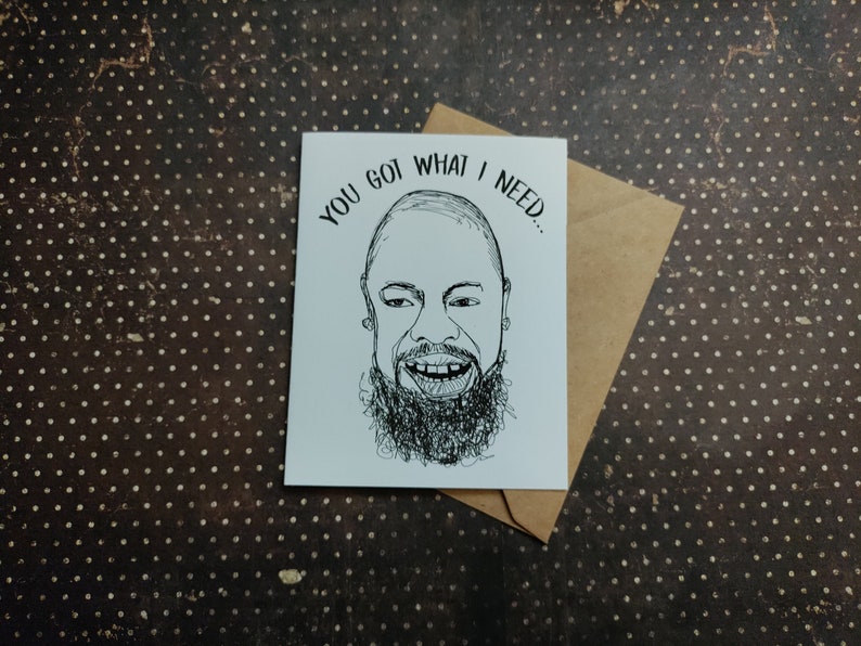 May include: A white greeting card with a black and white illustration of a smiling person with a beard. The card reads "YOU GOT WHAT I NEED..." and is placed on a brown envelope. The background is a brown surface with white polka dots.