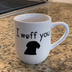 May include: White ceramic mug with a curved handle. The mug features the text "I woff you" above a black silhouette of a dog's head and paw prints. The mug is on a speckled countertop.