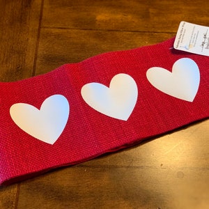 May include: Red burlap pillow wrap with three large white heart shapes. The pillow wrap has a tag that reads "Valentine Burlap Pillow Wrap" and includes the seller's name and contact information.