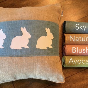 May include: A burlap pillow with a blue stripe and three white bunny silhouettes. The pillow is shown with rolls of burlap fabric in the colors sky, natural, blush, and avocado.
