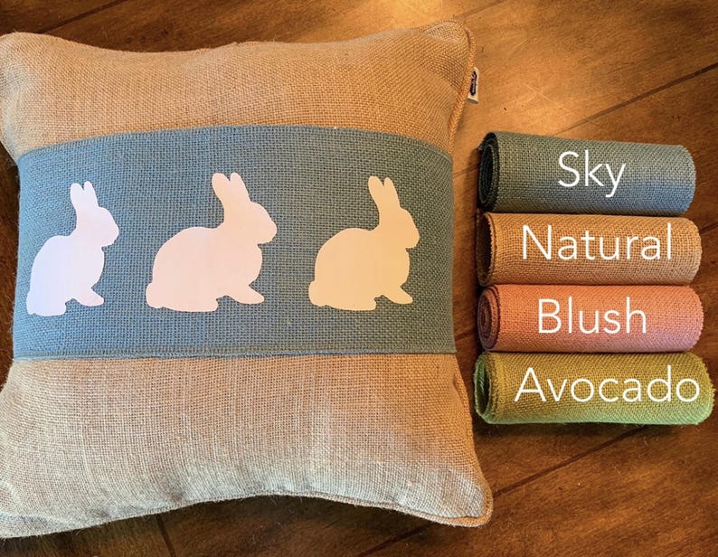 May include: A burlap pillow with a blue stripe and three white bunny silhouettes. The pillow is shown with rolls of burlap fabric in the colors sky, natural, blush, and avocado.