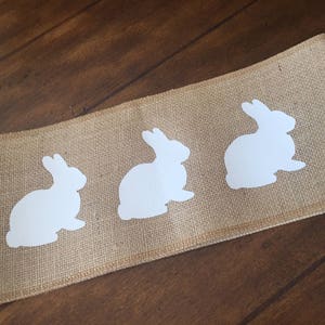 May include: A burlap table runner with three white bunny silhouettes. The runner is a natural brown color and the bunnies are evenly spaced.