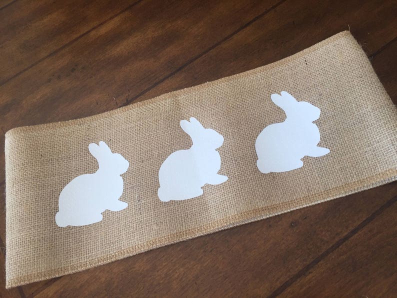 May include: A burlap table runner with three white bunny silhouettes. The runner is a natural brown color and the bunnies are evenly spaced.