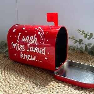 May include: Red metal mailbox with a white inscription that reads "I wish Miss Javaid knew..." with white stars and shooting stars on the side. The mailbox is open and the lid is laying on a woven surface.