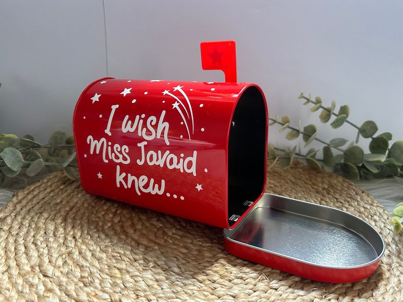 May include: Red metal mailbox with a white inscription that reads "I wish Miss Javaid knew..." with white stars and shooting stars on the side. The mailbox is open and the lid is laying on a woven surface.