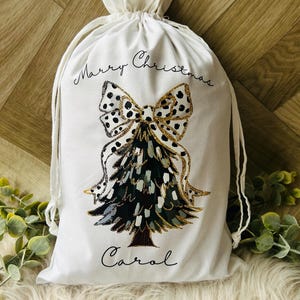 May include: White cotton gift bag with a black and white polka dot bow and a gold glitter Christmas tree design. The bag says "Merry Christmas Carol" in black script.