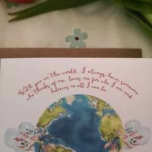 May include: A white card with a watercolor illustration of the Earth with green continents and blue oceans. The card has a pink floral design and the text "With you in the world. I always have someone who thinks of me, loves me for who I am and believes in all I can be."