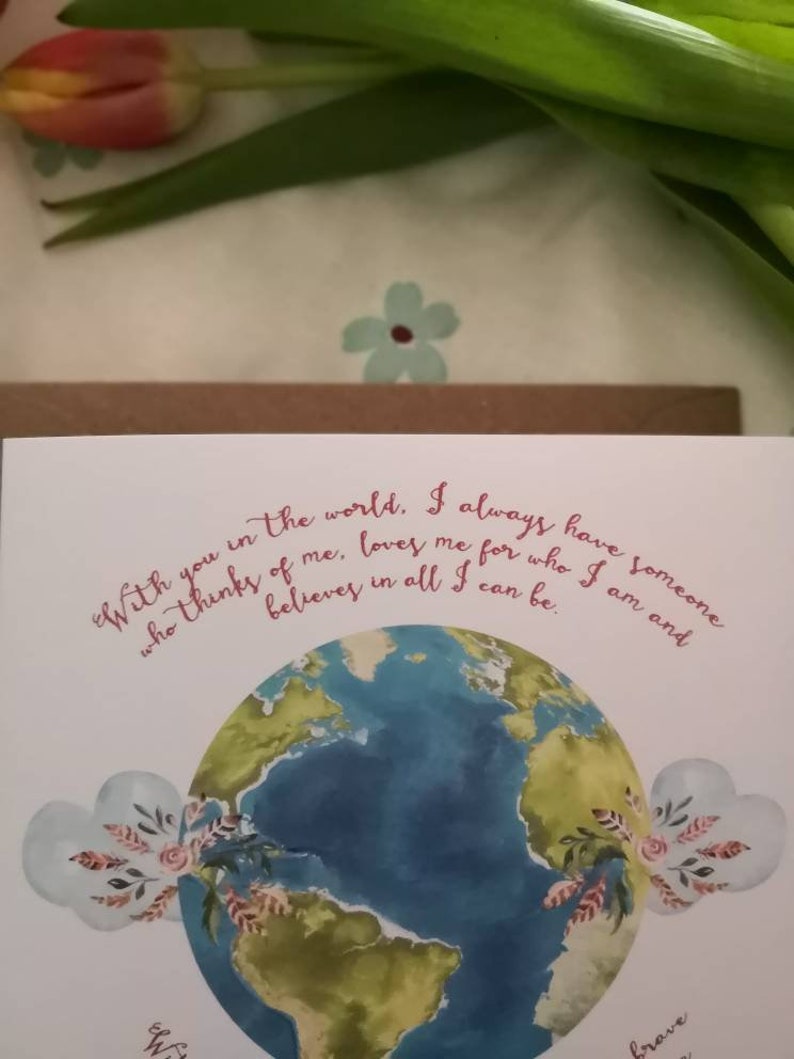 May include: A white card with a watercolor illustration of the Earth with green continents and blue oceans. The card has a pink floral design and the text "With you in the world. I always have someone who thinks of me, loves me for who I am and believes in all I can be."