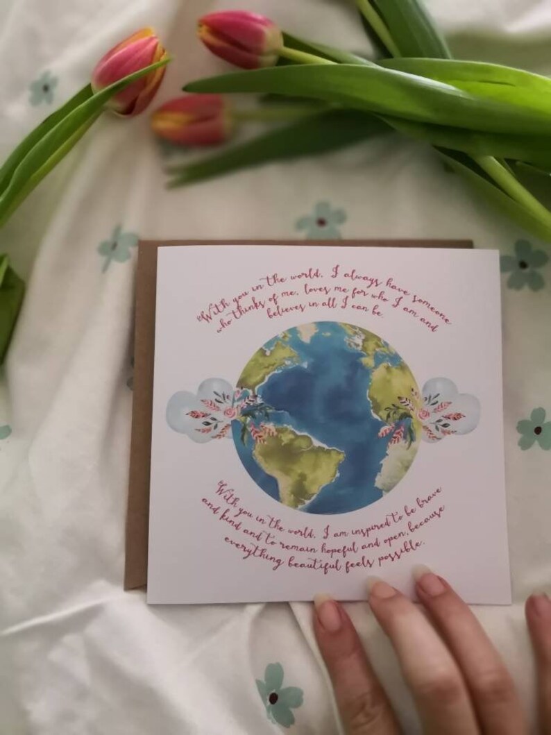 May include: A white card with a watercolor illustration of the Earth and pink flowers. The card has the text "With you in the world. I always have someone who thinks of me, loves me for who I am and believes in all I can be." and "With you in the world. I am inspired to be brave and kind and to remain hopeful and open. Everything beautiful feels possible."