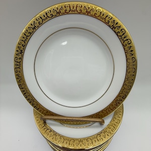 May include: A stack of ten white porcelain dessert plates with a gold rim and intricate gold scrollwork design.