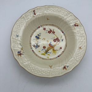 May include: A white ceramic bowl with a gold rim and a floral design featuring a bird and flowers. The bowl has a raised, textured rim.