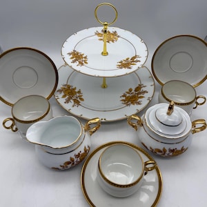 May include: A white tea set with gold accents and floral designs. The set includes a teapot, creamer, sugar bowl, four cups and saucers, and a two-tiered serving stand.