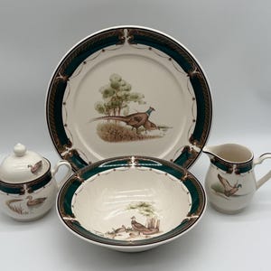 May include: A set of white ceramic dishes with a green and brown border featuring a pheasant and duck design. The set includes a large platter, a bowl, a creamer, and a sugar bowl.