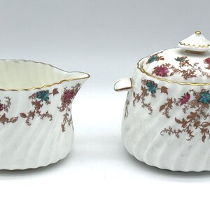 May include: A white creamer and sugar bowl set with a floral design. The set features a gold trim and a spiral design. The floral pattern includes teal and pink flowers with brown stems. The sugar bowl has a lid with a small finial.