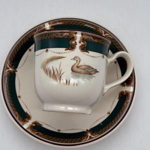May include: A white teacup and saucer set with a duck illustration. The cup and saucer have a dark green and brown border with an ornate design. The cup features a duck swimming in a pond with reeds.