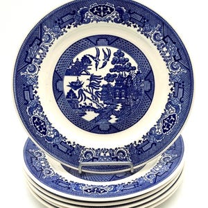 May include: A stack of blue and white Willow pattern plates. The plates feature a central landscape scene with birds and a detailed border design. The plates are stacked on a metal plate stand against a white background.