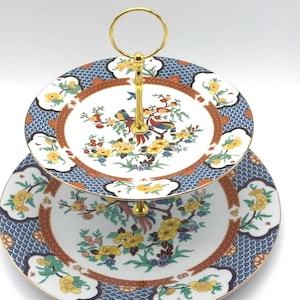 May include: Two-tiered serving tray with a gold handle. The plates feature a white center with a floral and bird design, surrounded by a brown border and a blue patterned edge. The tray is ideal for serving desserts or appetizers.