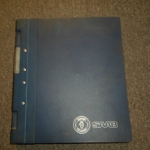 Puede incluir: Una carpeta azul con una columna vertebral de metal plateado y un logotipo Saab plateado con el texto "SAAB" impreso en blanco.