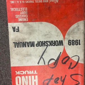 May include: A red and white Hino Truck Workshop Manual for the year 1989. The manual covers engine, chassis, body, and electrical components. The cover has the text "Shop Hino Truck" and "Copy" handwritten in black ink.