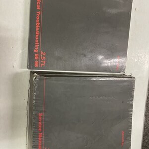 Op de afbeelding: Twee grijze autoreparatiehandleidingen met rode tekst. De bovenste handleiding heeft de titel "Elektrische probleemoplossing 2.5TL 95-96". De onderste handleiding heeft de titel "Servicehandleiding 2.5TL 95-96".