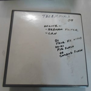 May include: A square, off-white object with black trim. Handwritten text in black ink includes "TELEMATICS 04", "ONLINE - HERMAN TESTER - CAN", and other technical terms. The object appears to be a data storage device.
