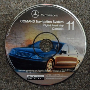 Puede incluir: Un CD plateado con un coche azul y marrón en la parte delantera. El texto en el CD dice "COMAND Navigation System Digital Road Map Canada 11".