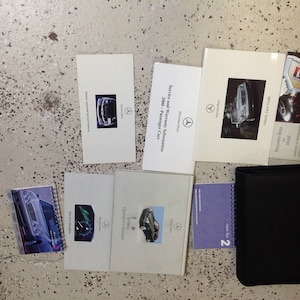 May include: A collection of car owner's manuals and service booklets for a Mercedes-Benz. The manuals are in a black binder with a silver Mercedes-Benz logo. The manuals include an operator's manual, service booklet, and vehicle care guide.