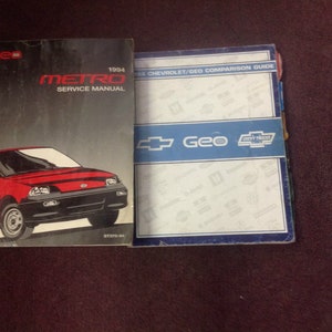 May include: Two automotive manuals are displayed. The first is a 1994 Geo Metro Service Manual, featuring a red car graphic. The second is a 1994 Chevrolet/Geo Comparison Guide, with blue and white accents and the Geo logo.