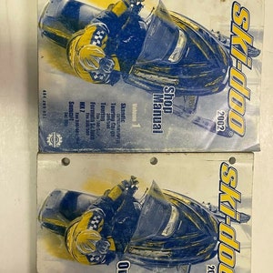 May include: Two manuals for a 2002 Ski-Doo snowmobile. The top manual is titled "Shop Manual" and the bottom manual is titled "One Day Book". Both manuals feature a yellow and blue snowmobile on the cover.