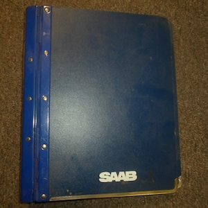 Puede incluir: Una carpeta azul con lomo azul y remaches metálicos. La carpeta tiene el logotipo blanco "SAAB" en la parte delantera. Las esquinas son doradas.