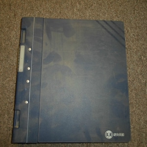 May include: A blue binder with a textured surface and a silver metal strip down the side. The binder has a silver metal logo that says "SAAB" on the bottom right corner.