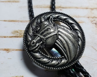 Puede incluir: Un bolo tie plateado con un diseño de cabeza de caballo. La cabeza del caballo está de perfil y tiene un diseño de cuerda alrededor. El bolo tie está en un cordón negro.