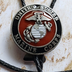 May include: A bolo tie featuring a circular emblem with the words "UNITED STATES MARINE CORPS." The emblem has a red center with a silver eagle, globe, and anchor design. The tie has a black braided cord.