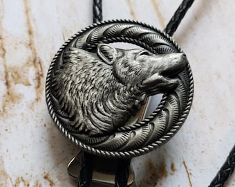 Puede incluir: Un bolo tie de plata con un diseño de cabeza de lobo. El lobo está mirando hacia la derecha y tiene un diseño detallado y texturizado. El bolo tie está hecho de cuero negro trenzado.