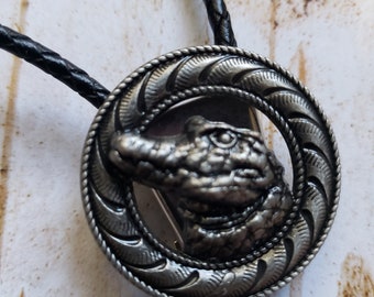 Puede incluir: Un bolo tie plateado con un cordón trenzado negro. El bolo tie presenta un colgante redondo plateado con un diseño de cabeza de cocodrilo. El colgante tiene una superficie texturizada detallada y un borde de cuerda.