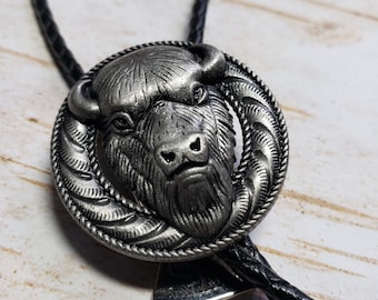 Puede incluir: Un corbatín bolo de plata con un diseño detallado de cabeza de búfalo. La corbata está hecha de cordón de cuero negro y tiene un cierre de plata.