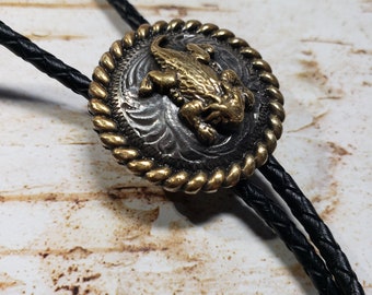 Puede incluir: Un corbatín de cuero negro con un colgante redondo de oro y plata con un diseño de lagarto.
