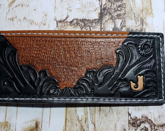May include: A handcrafted leather wallet with a detailed design. The wallet features a combination of black and brown leather, with intricate floral carvings and a textured pattern. A gold-colored letter "J" is visible.
