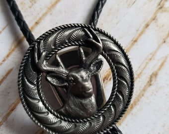 Puede incluir: Un bolo tie plateado con un colgante circular que presenta un diseño detallado de cabeza de ciervo. El colgante tiene un borde de cuerda y un fondo texturizado.