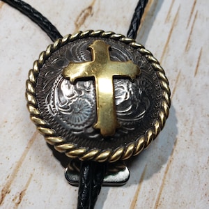 May include: A black bolo tie with a silver and gold round pendant featuring a gold cross design. The pendant has a rope-like border.