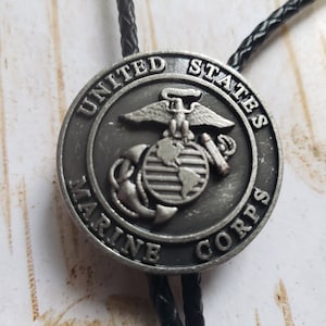 May include: A silver bolo tie with a round pendant featuring the United States Marine Corps emblem. The emblem depicts an eagle, globe, and anchor.