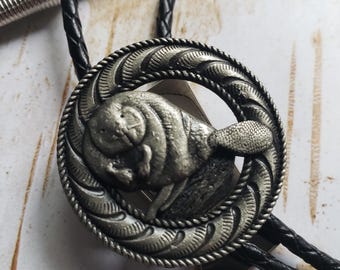 May include: A silver-toned bolo tie featuring a detailed, circular pendant with a central bison design. The pendant is encircled by a decorative rope-like border. The tie has a black braided leather cord and a silver-colored metal slide.